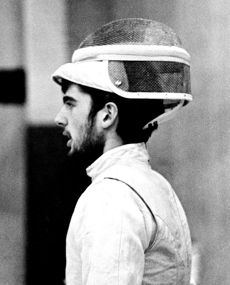 A black and white photo of Rocky as a younger man, in his fencing gear, with is mask up and balanced on the top of his head, looking away from the camera.